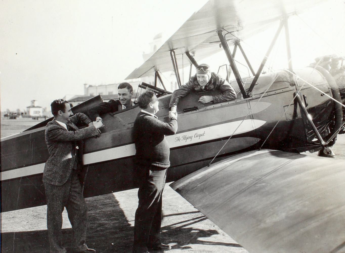 A black-and-white- photograph of two male pilots in an old airplane talking to two men standing on the tarmac.