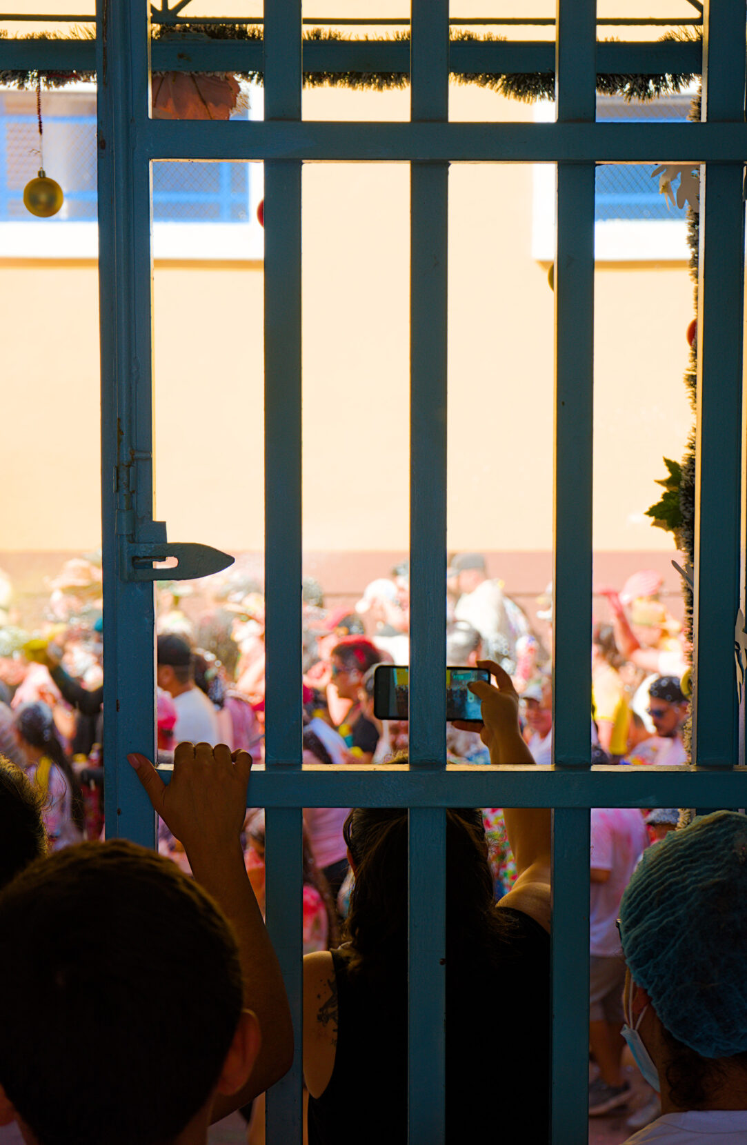 A spectator records a colorful parade on her phone behind a barred gate.
