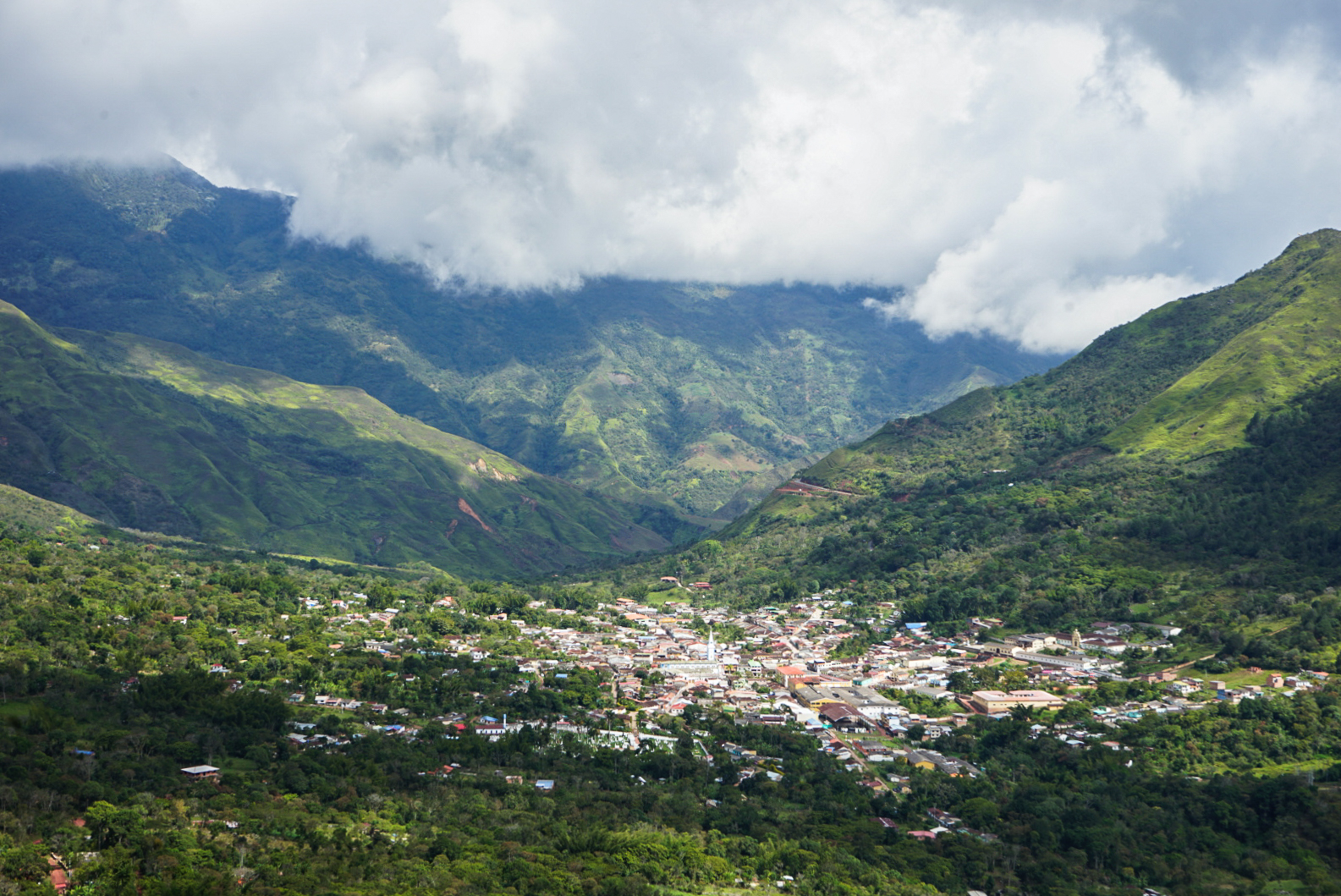 A small town sits in a lush forested valley surrounded by mountains and clouds.
