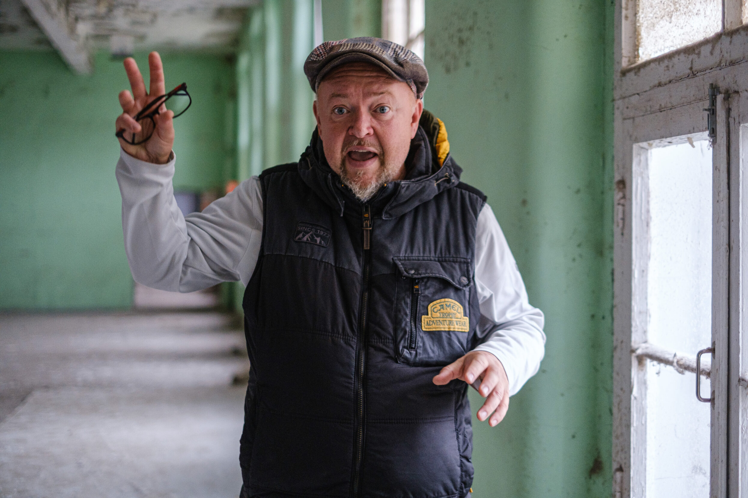 A man wearing a hat and vest gestures with outstretched hands in an empty concrete building.