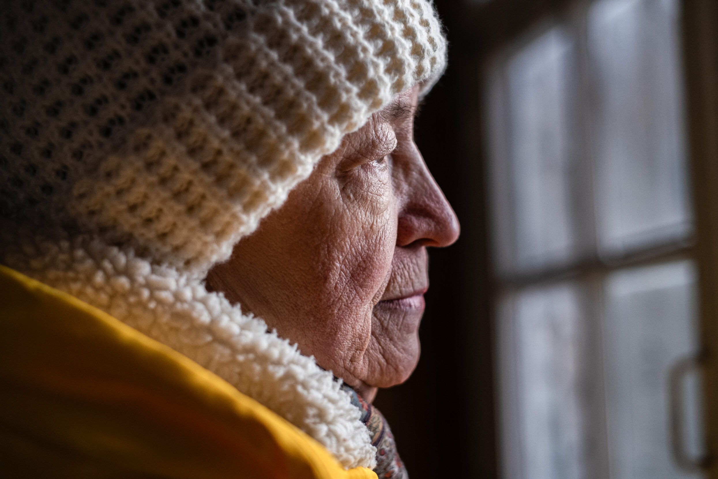 An older woman in a woolen white hat and coat gazes toward naturally lighted windows.