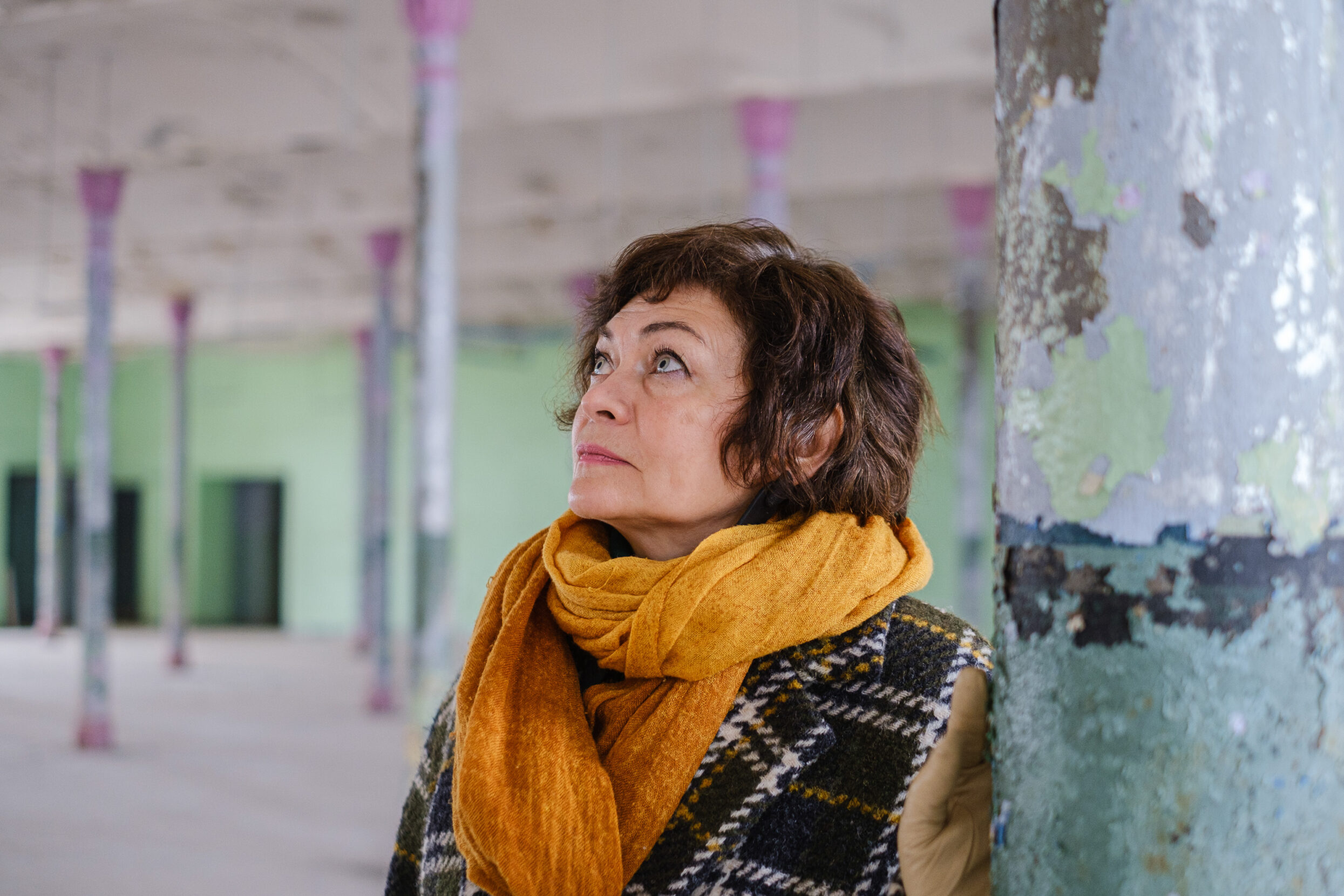 A woman in a yellow scarf with short brown hair and gray eyes stands amidst old columns with peeling paint.