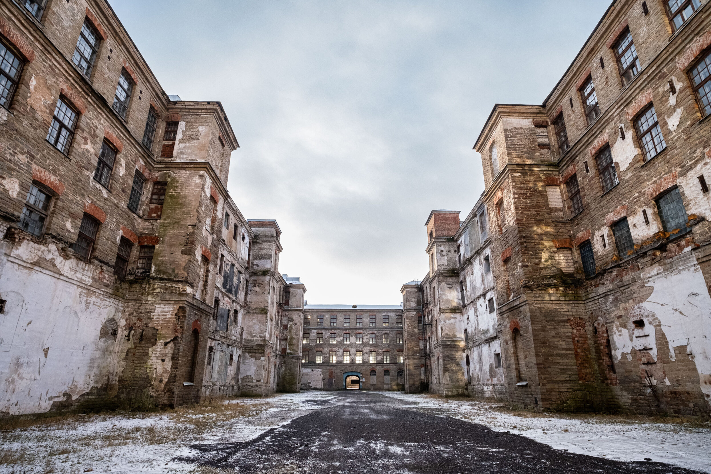 An old snowy paved road flanked by tall stone and brick abandoned buildings.