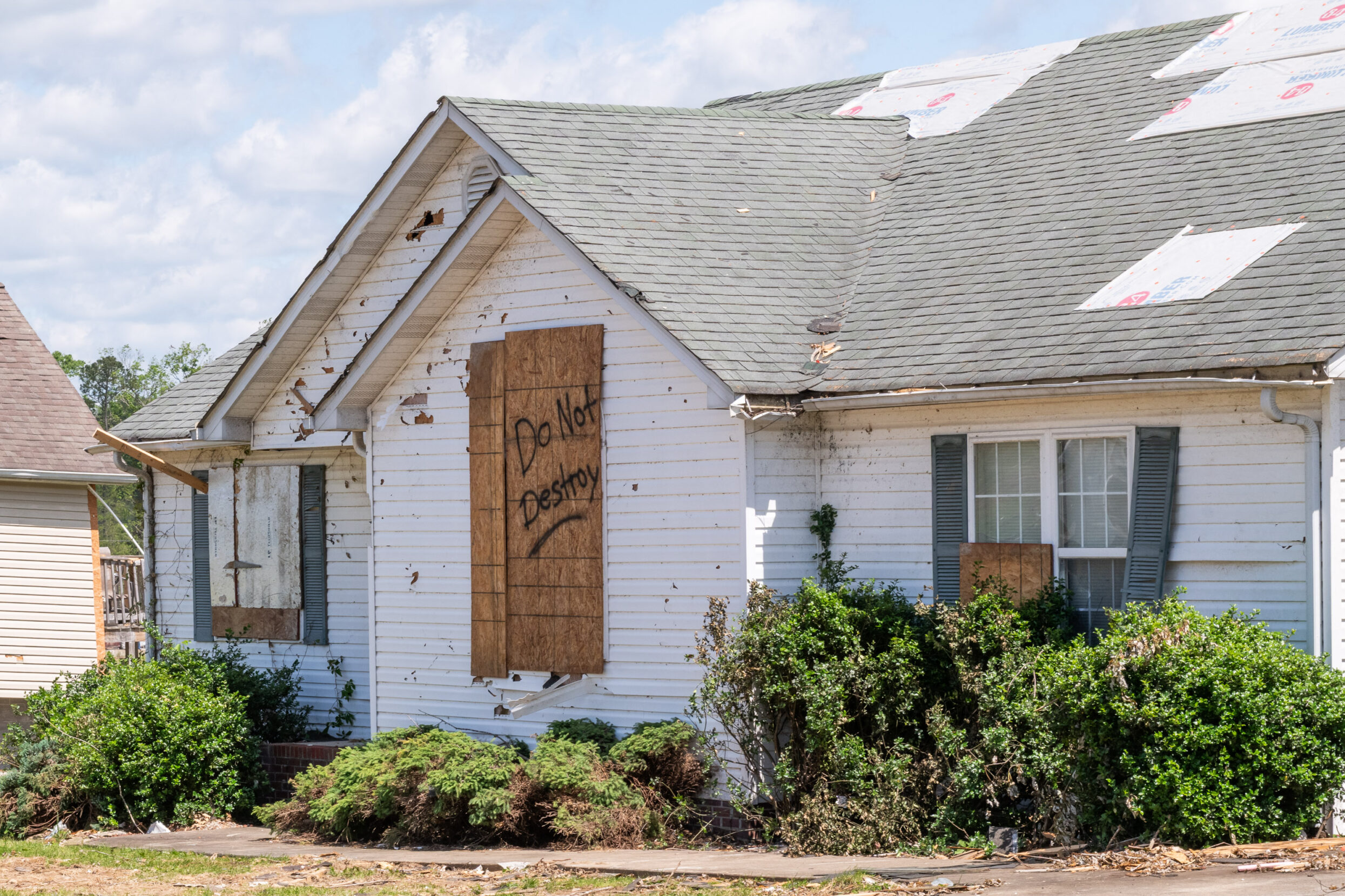 A house with tarps on the roof and a boarded up window with the words Do Not Destry spray-painted on the boards.