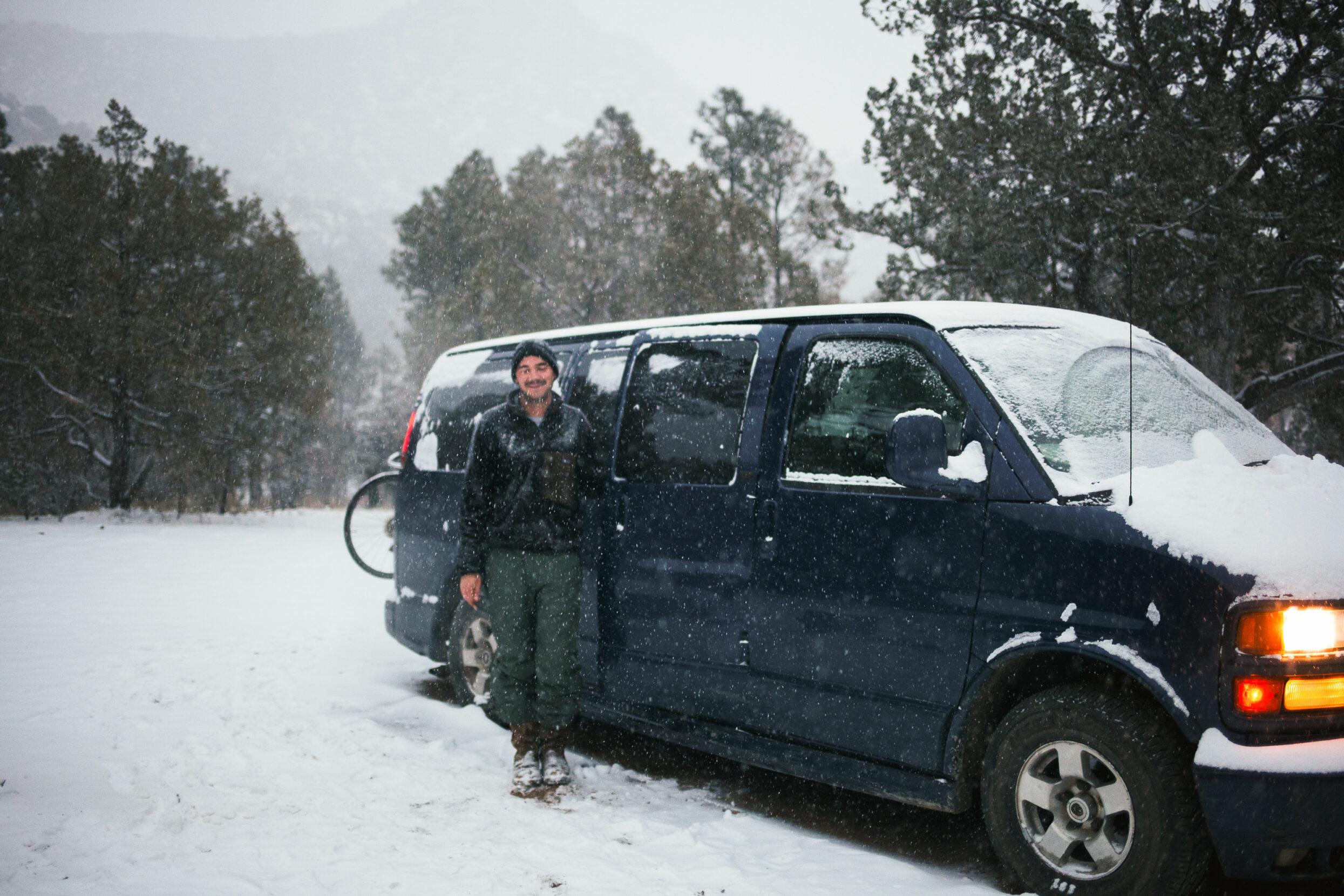 A man in a beanie and jacket stands before a blue van in the falling snow.