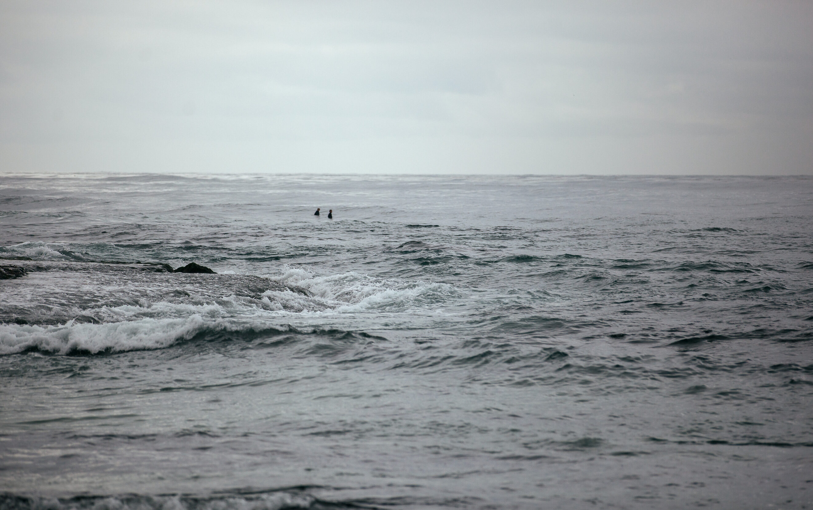 Two surfers float between waves below an overcast sky.