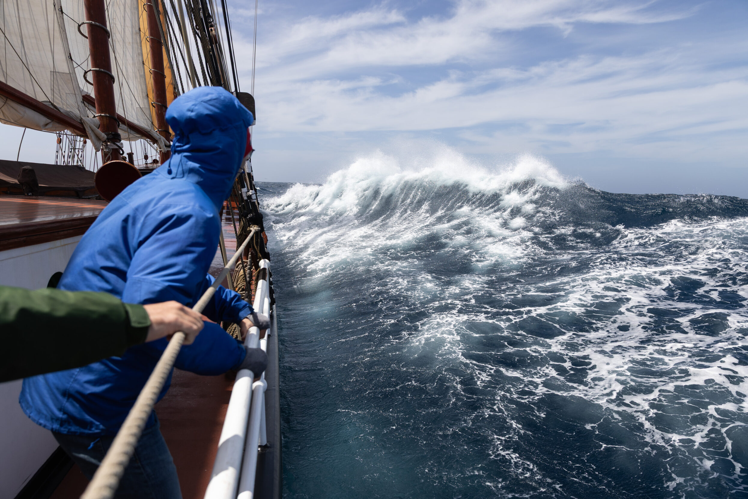 A sailer grasps the edge of their wooden boat and looks out to rolling waves.