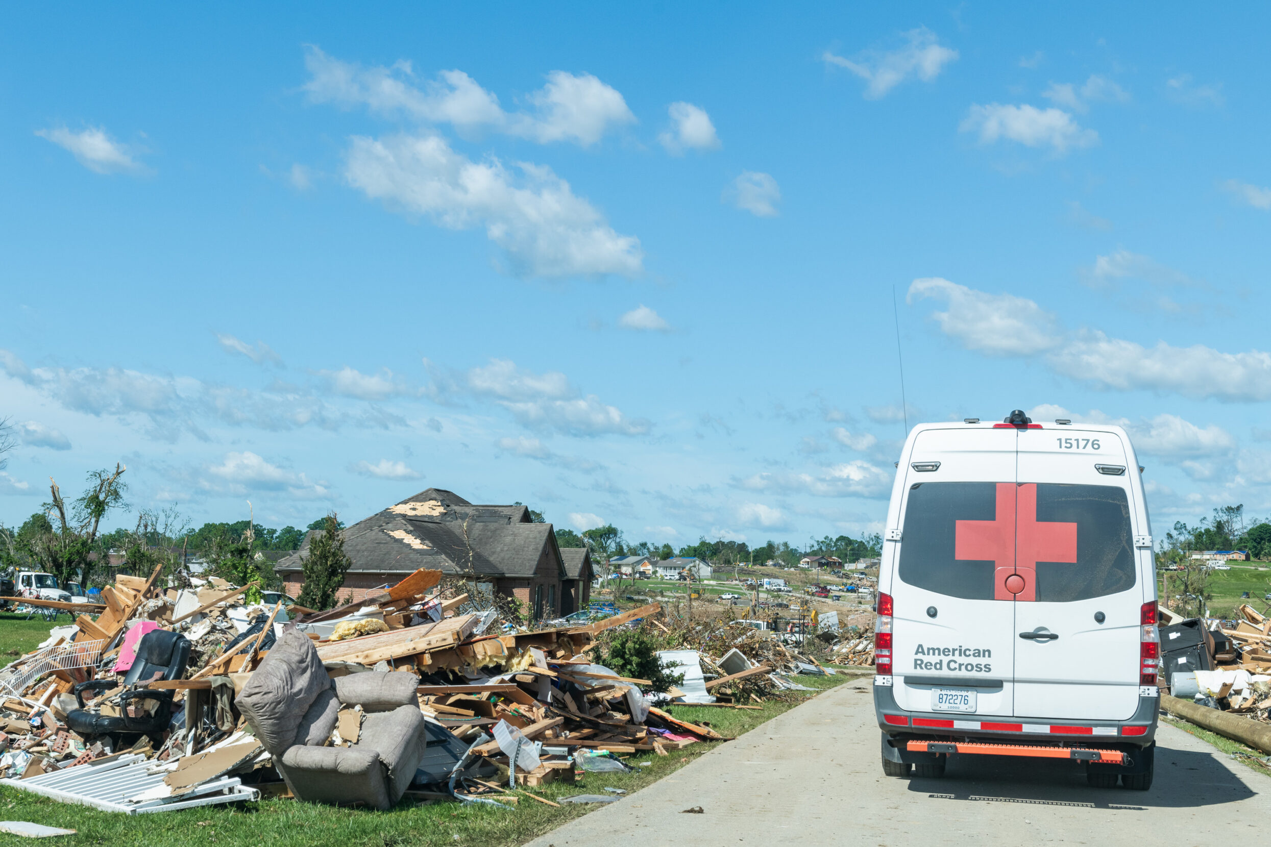 Debris from a tornado lines a street under a blue sky while a disaster response vehicles drives down the street.