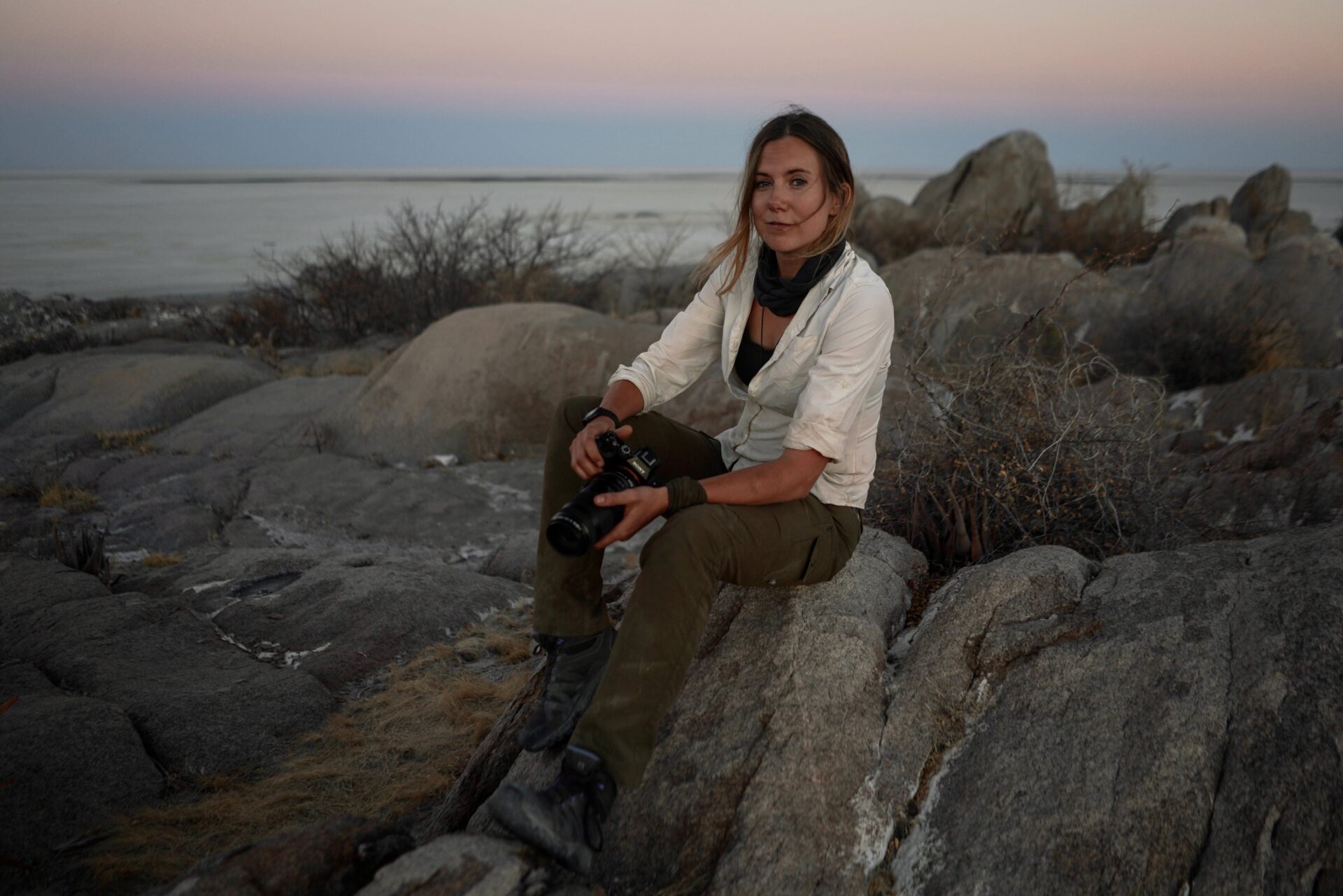 A woman holding a camera sits on a rock by the coast at sunset.