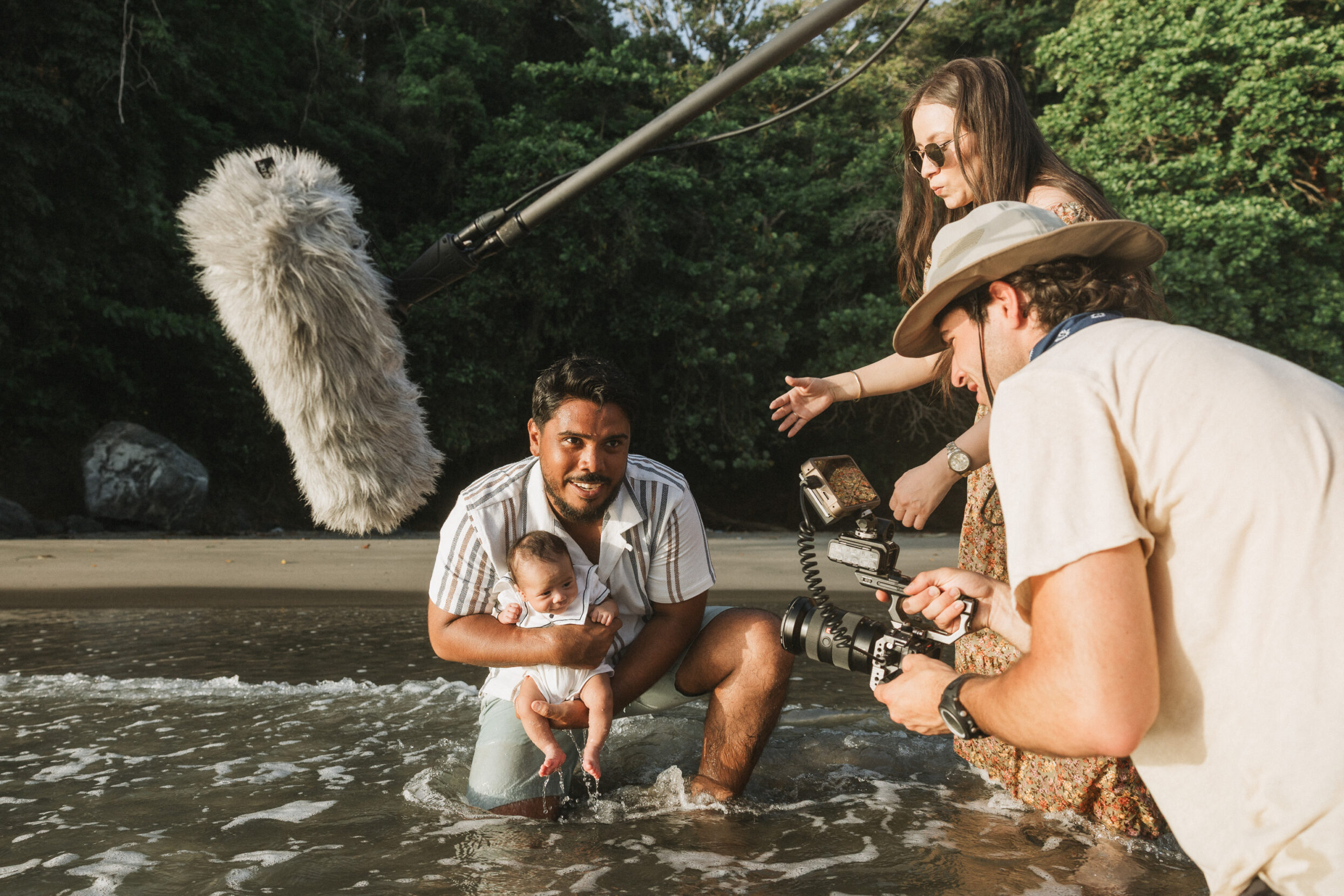 A behind-the-scenes image of a man holding a baby in the water while a man and woman film him