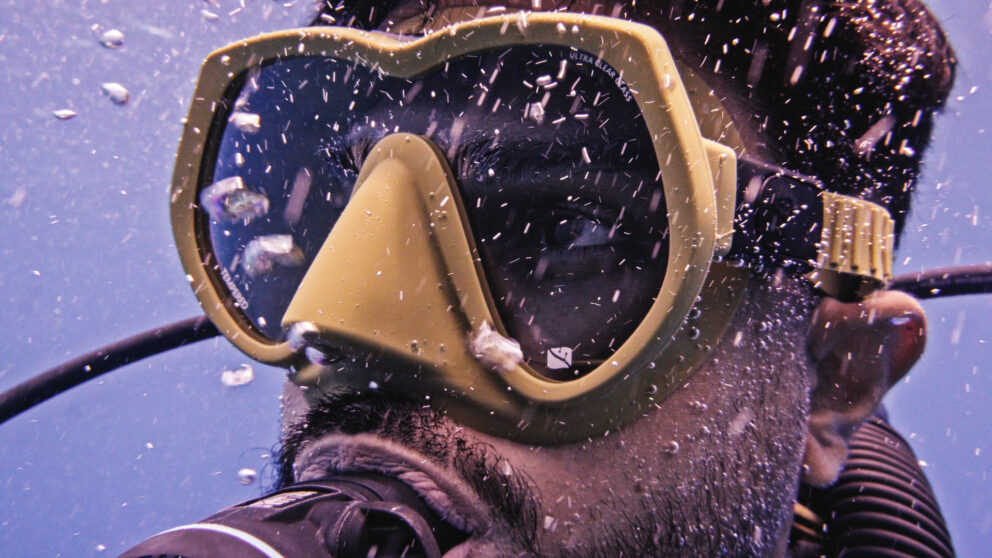 A closeup of a man with a dive mask on underwater with bubbles around him