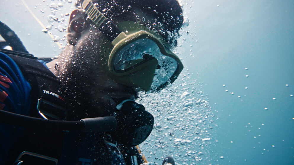 A closeup of a man with a dive mask on underwater with bubbles around him
