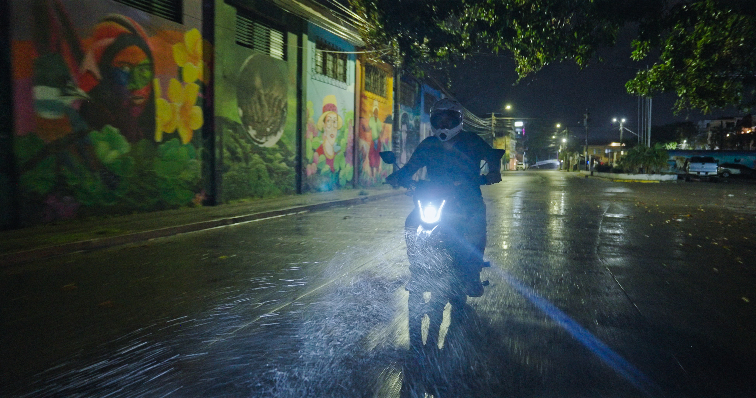 a man on a motorcycle in the rain at night with colorful murals alongside him