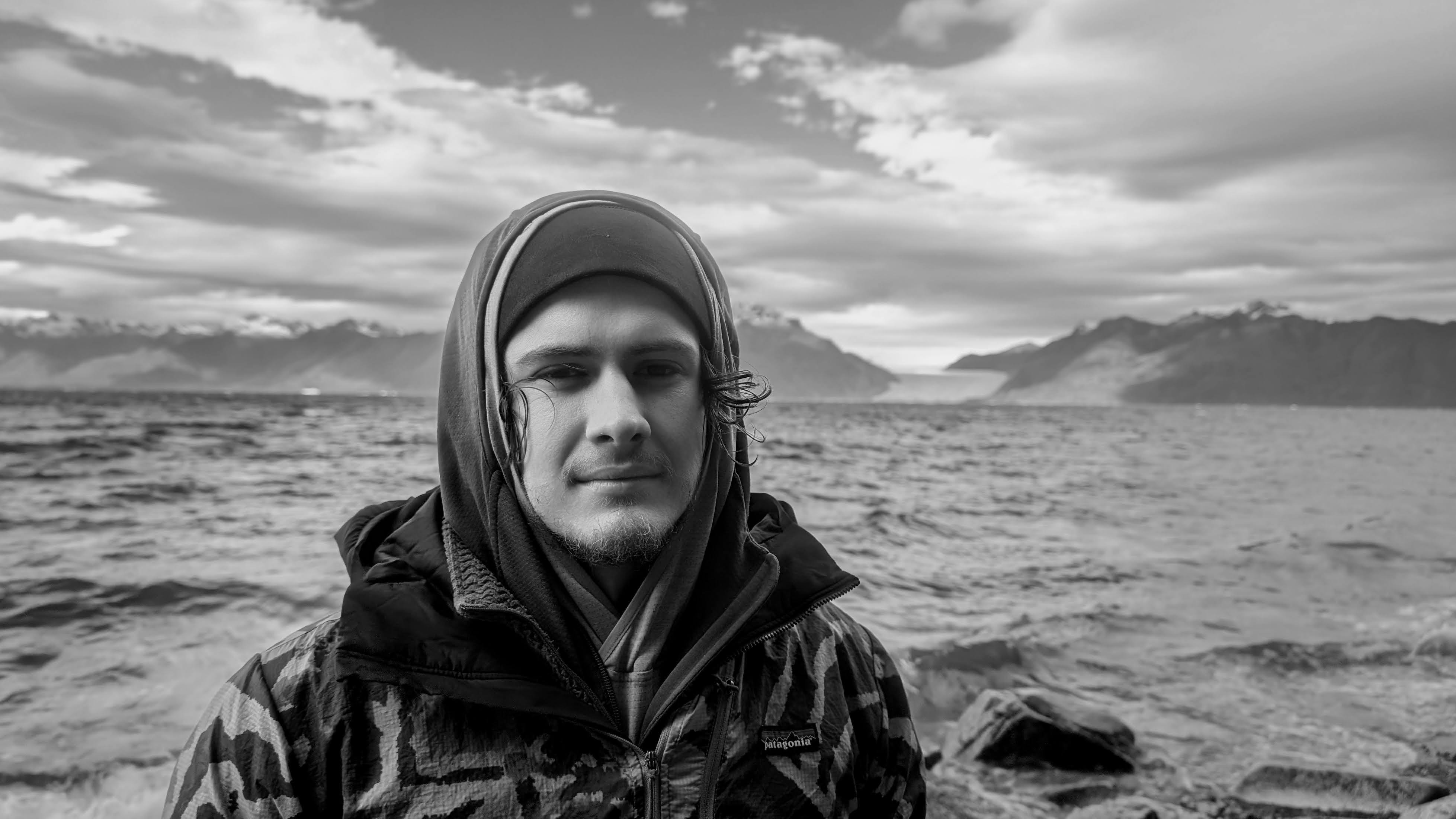 Black and white portrait of a man in a jacket and beanie with a large body of water and mountains in the background.