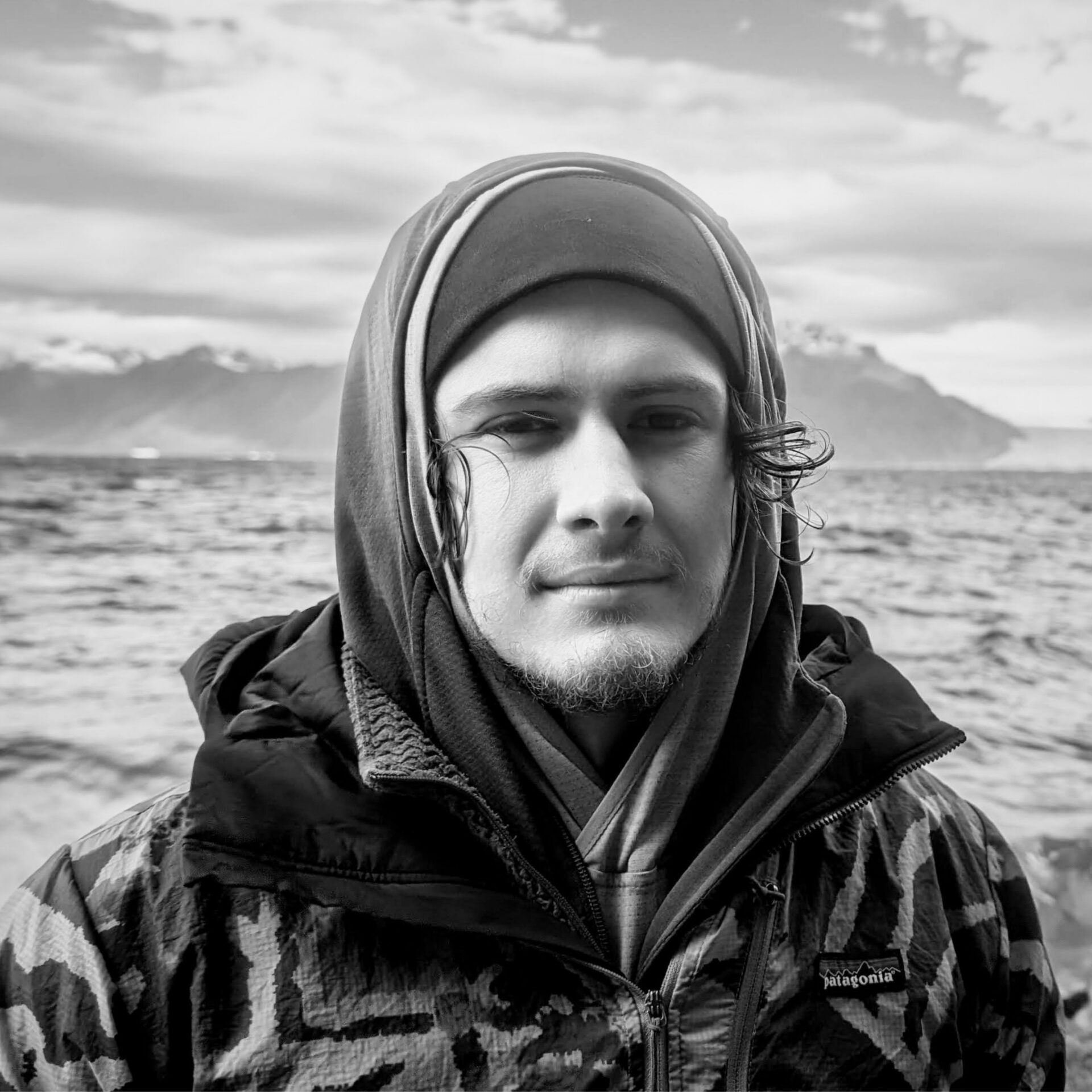 A man in a beanie and hood with mountain and the oceans behind him.